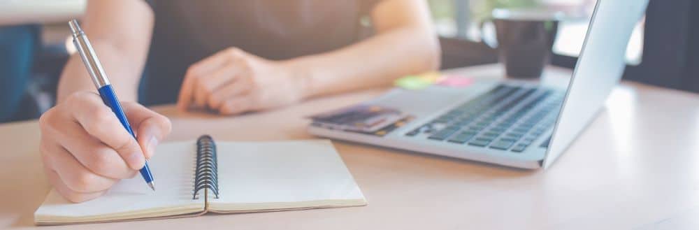 Nainen kirjoittaa muistikirjaan, vieressä pöydällä läppäri.